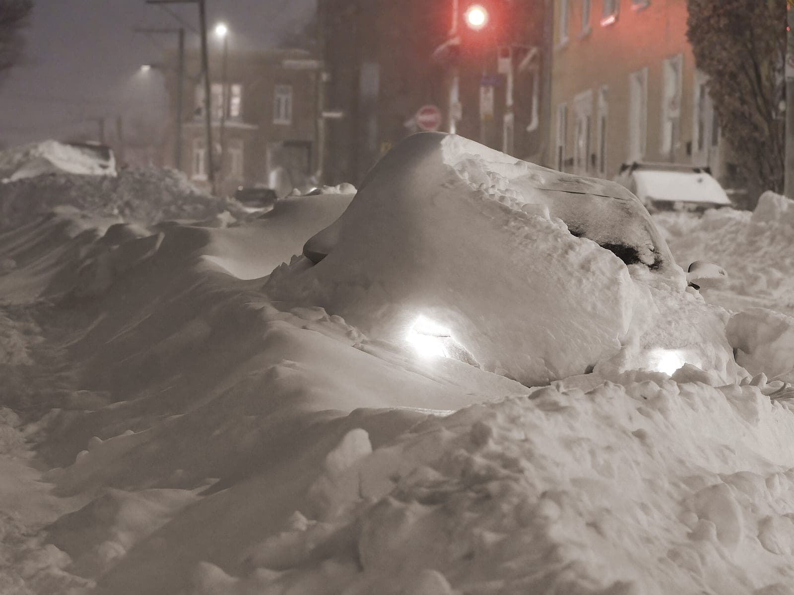 Rue enneigée la nuit après une bordée : une voiture ensevelie, phares allumés, entre deux bancs de neige