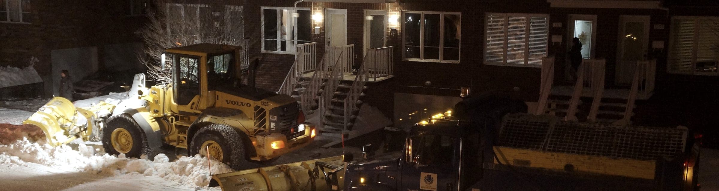 Opération de déneigement de nuit : chargeuse et camion à lame, gyrophares allumés, devant des habitations