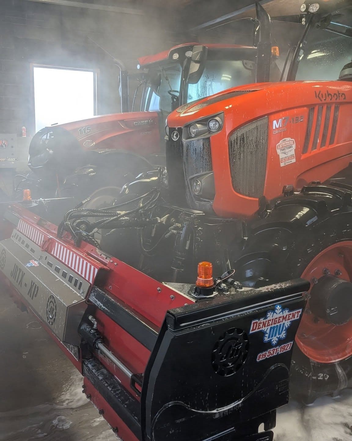 Chasse-neige JRTX-XP attelé à un tracteur Kubota dans le garage, le logo de Déneigement MV peint sur le côté de la lame