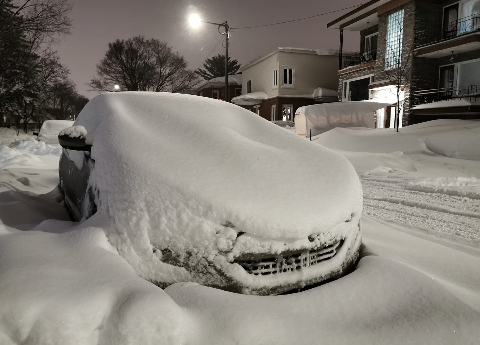 Voiture ensevelie sous la neige devant des maisons, la nuit, après une bordée